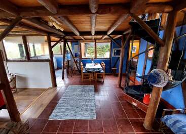 This cozy cabin interior features wooden beams, blue walls, red tiles, a spiral staircase, rustic furniture, a dining table, large windows, and lots of natural light.