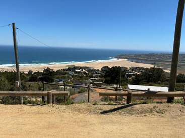 This image shows a coastal view with a sandy beach, blue ocean, clear sky, scattered houses, green trees, wooden fences, and utility poles under bright sunlight.