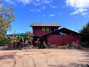 A rustic two-story red house with a sloped roof stands under a blue sky, surrounded by greenery, potted plants, and a dirt driveway in front.
