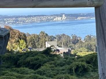 A beautiful coastal landscape featuring green bushes, trees, and houses in the foreground, a vast blue ocean, distant buildings, and hills under a clear sky.