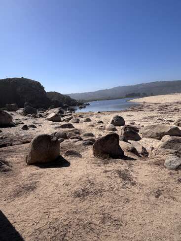 Rocky sand beach under a clear blue sky. Calm water is surrounded by large boulders, distant hills, and a serene, natural landscape. Peaceful and sunny.