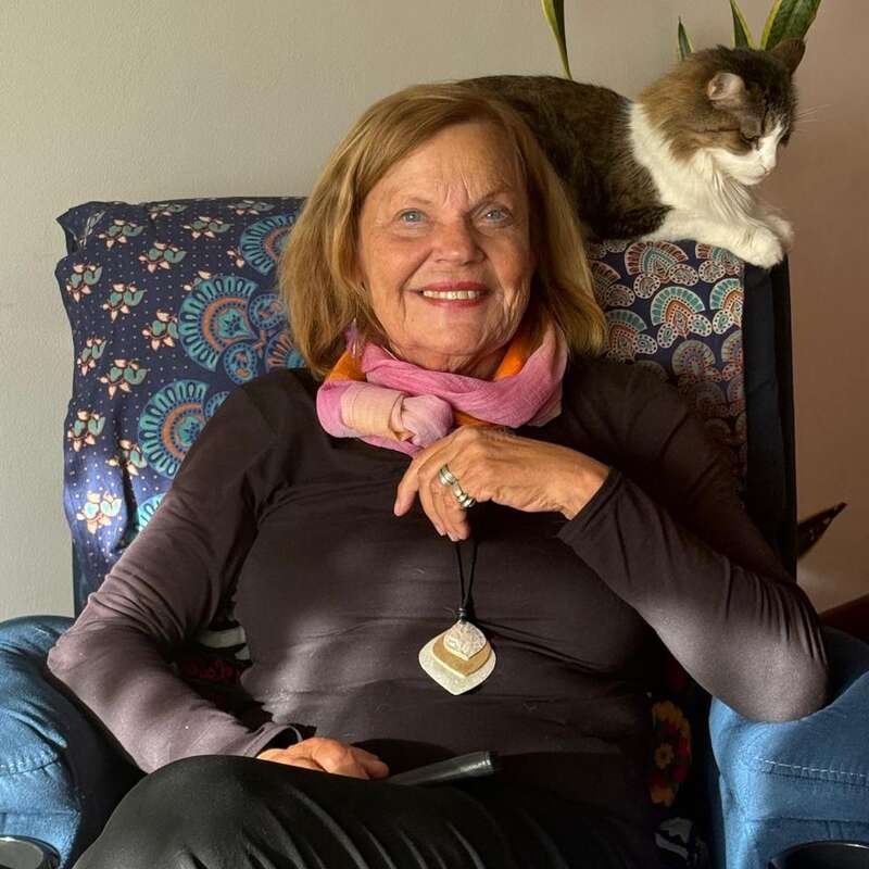 A smiling older woman sits in a patterned armchair, wearing a pink scarf and a pendant necklace, with a content cat perched on the chair’s back.
