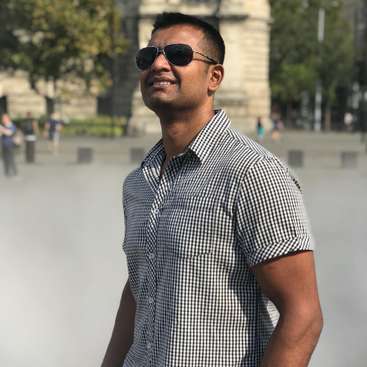 The image depicts a man standing outdoors, wearing sunglasses and a short-sleeved checkered shirt, with a blurred background of trees and buildings.