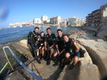 Cinco personas con equipo de submarinismo están sentadas en una orilla rocosa, sonriendo y posando. Detrás de ellos, el mar y los edificios de la ciudad crean un ambiente animado y veraniego.