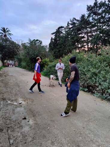 Tres hombres y un perro se encuentran en un camino de tierra rodeado de vegetación y árboles altos, mientras se ve a dos personas caminando a lo lejos.