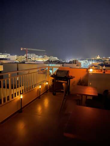 Una acogedora terraza en la azotea por la noche, suavemente iluminada por luces cálidas. Mesas y sillas frente a un paisaje urbano con edificios iluminados, una grúa y vistas lejanas del puerto.