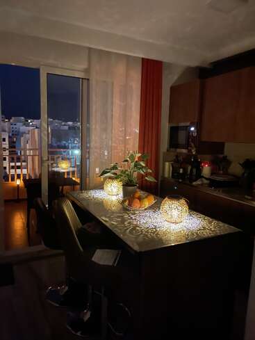 Una acogedora cocina nocturna con farolillos de bellos motivos que proyectan intrincadas sombras sobre la encimera, un frutero, una maceta y una atractiva vista desde el balcón.