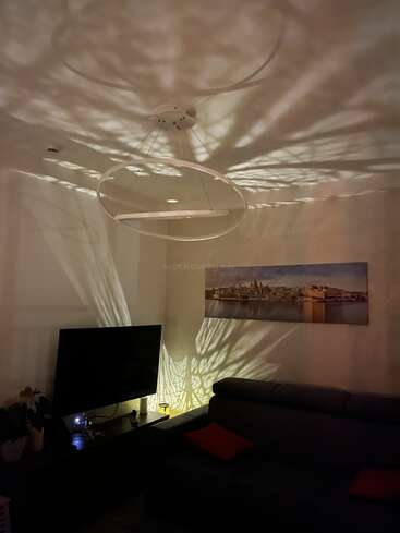 Un moderno salón presenta llamativos patrones de luz en el techo y la pared, proyectados por una lámpara de techo decorativa, sobre un sofá, un televisor y obras de arte en la pared.