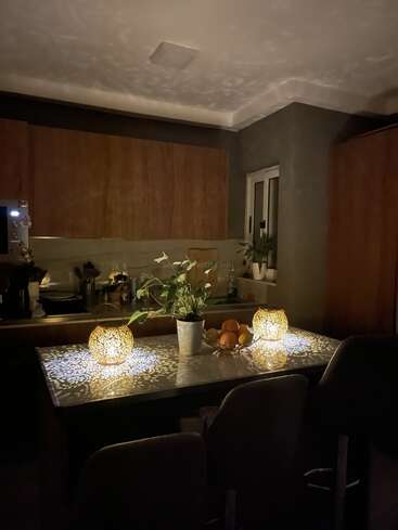 Una acogedora cocina nocturna con una cálida iluminación. Unas lámparas ornamentadas proyectan sombras intrincadas sobre la mesa, que sostiene una planta y un cuenco con frutas variadas.