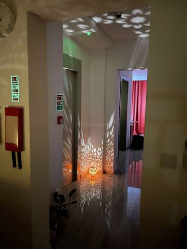 Un pasillo cálidamente iluminado presenta intrincados patrones de luz proyectados por una lámpara decorativa en el suelo. Los reflejos bailan sobre el suelo brillante, creando una atmósfera acogedora y acogedora.