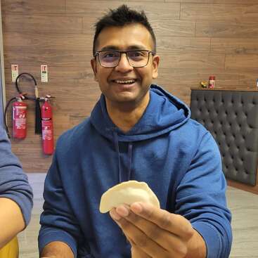 Un hombre sonriente con gafas y sudadera azul con capucha sostiene un trozo de comida. Detrás de él hay extintores, un banco acolchado y una pared de madera.