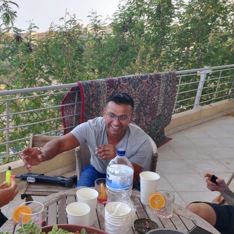 Un hombre sonriente sentado en un patio con amigos, rodeado de bebidas, tazas, un cenicero y una gran botella de agua. Árboles y una alfombra al fondo.