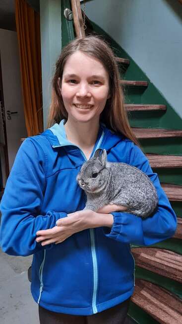 A smiling woman in a blue jacket stands indoors, holding a fluffy gray rabbit in her arms. She stands by a staircase with wooden steps behind her.