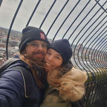 A happy couple, warmly dressed in jackets and beanies, embraces and smiles for a selfie on a high, fenced viewpoint overlooking a cityscape on a cloudy day.