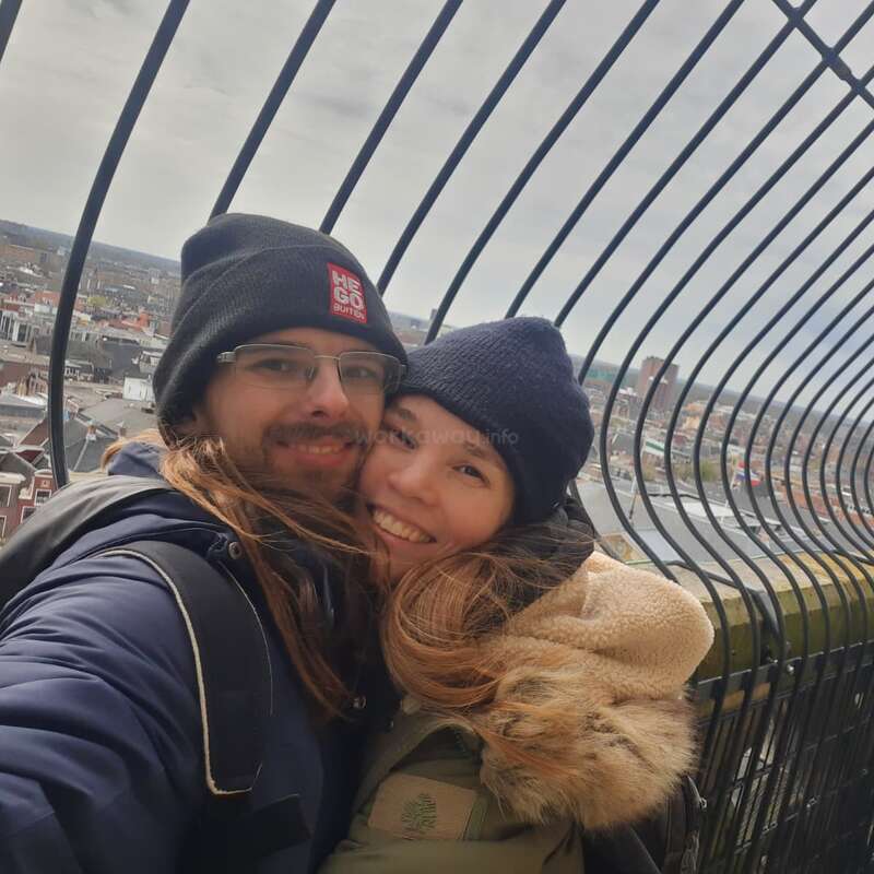 A happy couple, warmly dressed in jackets and beanies, embraces and smiles for a selfie on a high, fenced viewpoint overlooking a cityscape on a cloudy day.