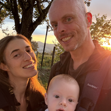 A happy family of three poses outdoors at sunset. The parents smile warmly, holding their baby close. Trees and hills are visible in the glowing background.