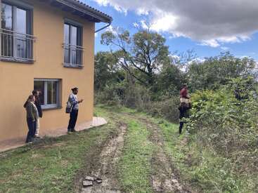 Four people stand beside a yellow house, observing two others near bushes. A dirt path curves through green grass, with trees and a cloudy blue sky overhead.