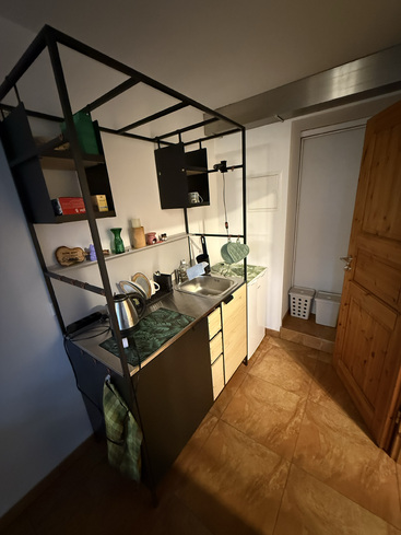 This image shows a compact kitchenette with open shelving, a small sink, countertop, kettle, various kitchen utensils, green décor, tiled floor, and a wooden door.