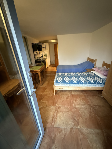 A small cozy room with a bed, blue blanket, and pillows. There’s a table, chairs, open shelves, and visible kitchen area near the entrance.