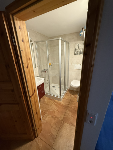 This image shows a small, modern bathroom with beige tiles, a glass shower, a toilet, a small sink with a cabinet, wooden door frame, and a wall picture.