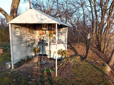 A small outdoor shrine with religious icons and flowers stands in a forest clearing. The structure is simple, surrounded by bare trees and sunlight. Peaceful atmosphere.