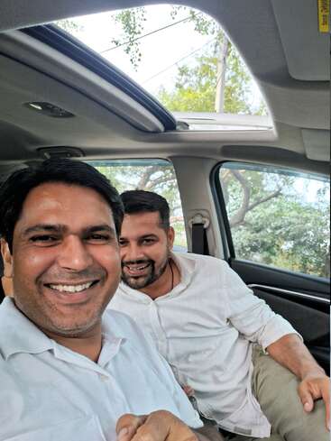 Two men in white shirts are smiling and taking a selfie inside a car with an open sunroof. Trees and daylight are visible outside the windows.