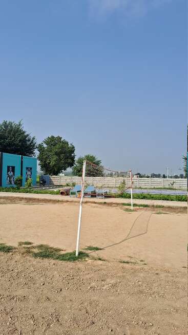 A sandy volleyball court with a net stands under a clear blue sky, surrounded by trees, a fence, benches, and a blue building featuring wall art.