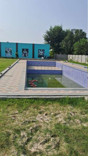 An empty, partially filled swimming pool with murky water sits outdoors beside a blue building adorned with bodybuilder posters, surrounded by green grass and trees.