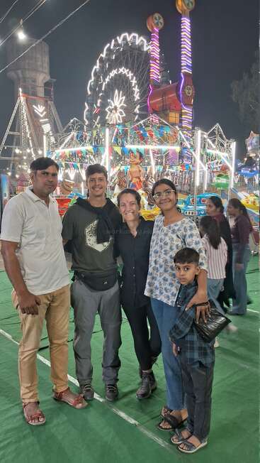 A group of five people pose at a lively carnival at night. Bright lights, rides, and a Ferris wheel create a festive and joyful background atmosphere.