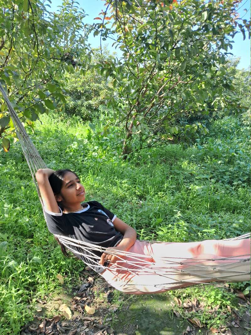 A young girl with dark hair and a black shirt lies in a white hammock, surrounded by lush greenery and trees, basking in the sunlight.