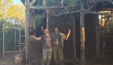 Zwei Menschen stehen lächelnd mit erhobenen Armen vor einem rustikalen, teilweise gebauten Gebäude aus Holz und natürlichen Materialien, umgeben von Bäumen und Sonnenlicht.