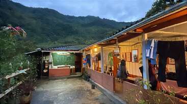 Una acogedora casa rural al atardecer, enclavada en exuberantes montañas verdes. La ropa tendida cuelga en el exterior, las cálidas luces brillan y las plantas bordean el patio. Un ambiente tranquilo, rústico y acogedor.