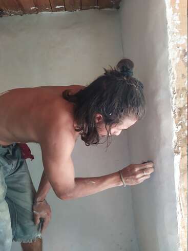 Eine Person ohne Hemd, mit langen Haaren, die zu einem Knoten zusammengebunden sind, schleift oder glättet eine weiße Wandecke; sie trägt Shorts und befindet sich in einem Raum mit Holzdecke.