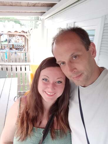 Un couple souriant pose de près pour un selfie sous un porche couvert, profitant d'une journée ensoleillée. Des sièges extérieurs et une structure en bois sont visibles derrière eux.