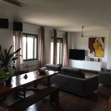 L'image représente un salon avec une table à manger, un canapé, un téléviseur et des œuvres d'art, avec des murs blancs, des planchers en bois foncé et de grandes fenêtres avec des rideaux beiges.