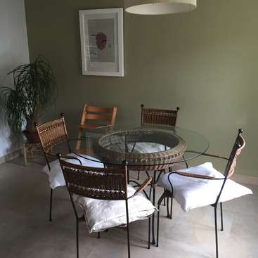 L'image représente une salle à manger avec une table ronde en verre et quatre chaises, avec une plante et des œuvres d'art au mur.