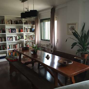 L'image représente une salle à manger confortable avec une table en bois, des étagères et une fenêtre, dans une atmosphère chaleureuse et accueillante.
