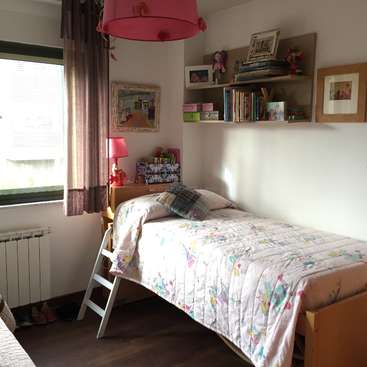 L'image représente une petite chambre confortable avec un lit simple, une fenêtre et une étagère avec des livres et des jouets. La pièce est bien éclairée et accueillante.