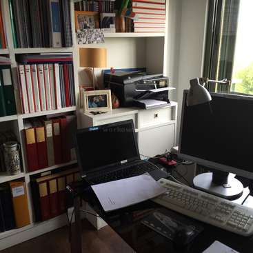 L'image représente un bureau à domicile avec un bureau, un ordinateur portable et une étagère, avec une imprimante, une lampe et une fenêtre avec vue sur les arbres à l'extérieur.