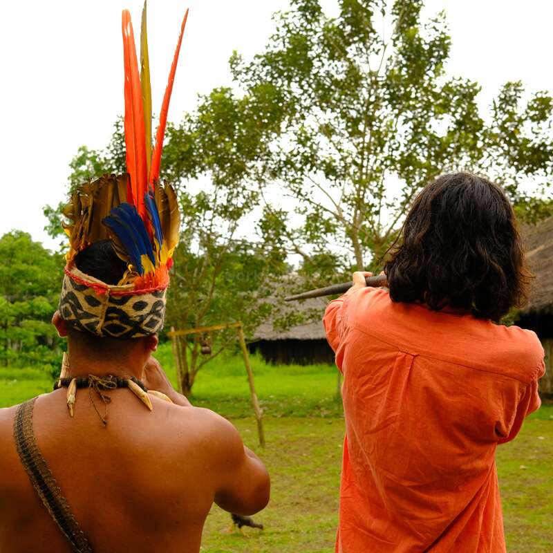 Two people stand outdoors. One wears a colorful feathered headdress and tribal jewelry, while the other, in an orange shirt, aims a blowgun at a target.