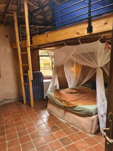 Esta acogedora habitación rústica cuenta con una cama redonda con mosquitera, suelos de baldosas de terracota, un altillo de madera con escalera, iluminación natural y decoración terrosa.