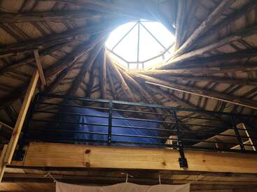 Una acogedora zona de loft con una barandilla rústica de madera se encuentra bajo una claraboya circular, rodeada de vigas de madera arremolinadas, creando un refugio cálido, acogedor e inspirado en la naturaleza.