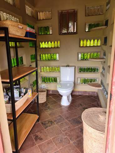 Esta imagen muestra un singular cuarto de baño con inodoro, estanterías con artículos de aseo y paredes hechas con botellas de vidrio. Las cestas tejidas y los suelos de baldosas añaden un toque rústico.