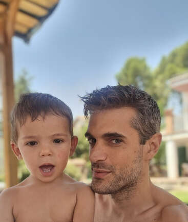 Um homem sem camisa e um menino posam bem próximos um do outro ao ar livre, ambos olhando para a câmera. No fundo, há árvores, prédios e um céu azul claro.