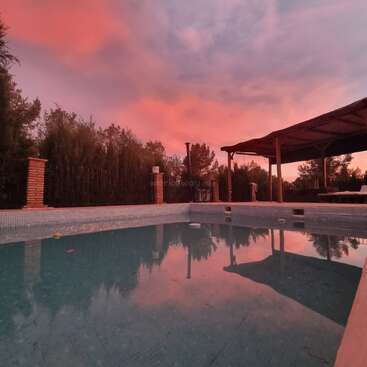 Una piscina vacía refleja un espectacular cielo anaranjado y púrpura al atardecer, rodeada de árboles y una pérgola de madera que ofrece sombra, en un tranquilo entorno al aire libre.