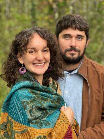 Une femme souriante aux cheveux bouclés et aux boucles d'oreilles violettes se tient à côté d'un homme vêtu d'une veste marron. Ils sont en plein air, entourés de verdure, et ont l'air satisfaits.