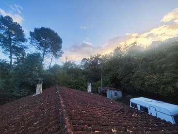 Un toit rustique en tuiles rouges surplombe une forêt dense au coucher du soleil. Une caravane blanche et un petit hangar se trouvent à proximité, tandis que les silhouettes des grands arbres se découpent sur le ciel.