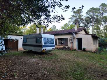 Une petite maison rustique au toit de tuiles rouges se trouve dans une cour boisée. Une vieille caravane est garée devant, entourée d'arbres et de feuilles éparses.