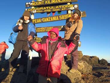 Drei Bergsteiger in Winterausrüstung posieren triumphierend am Gipfelschild des Kilimandscharo und feiern ihre Leistung unter blauem Himmel, umgeben von felsigem Gelände und Sonnenlicht.