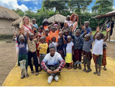 Das Bild zeigt eine Gruppe von Kindern und zwei Erwachsenen, die für ein Foto auf einer gelben Matte in einer dörflichen Umgebung posieren, mit strohgedeckten Hütten und Bäumen im Hintergrund.